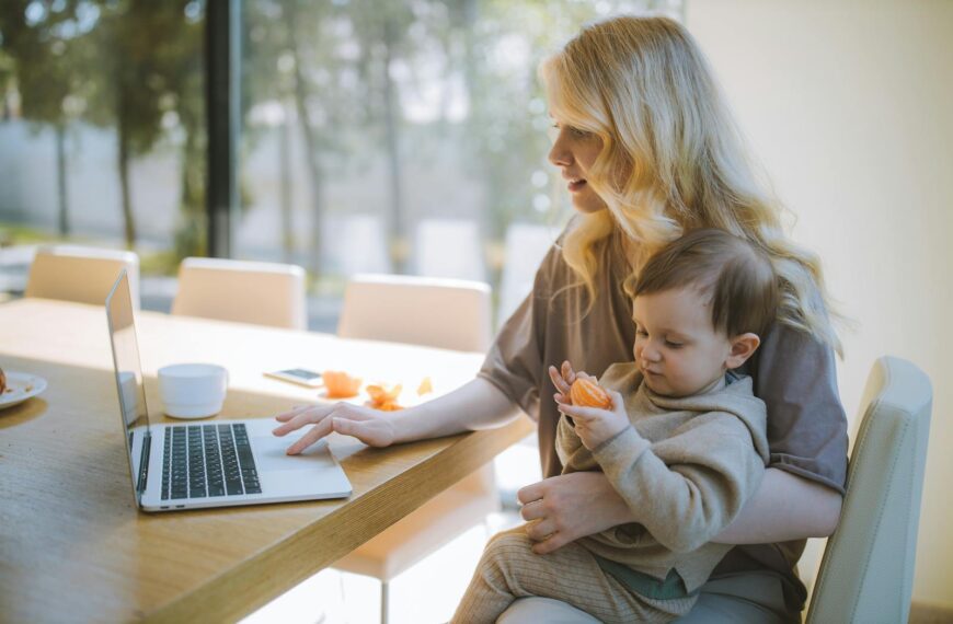 A mother works on a laptop at home while holding her baby, showcasing remote working and parenting.