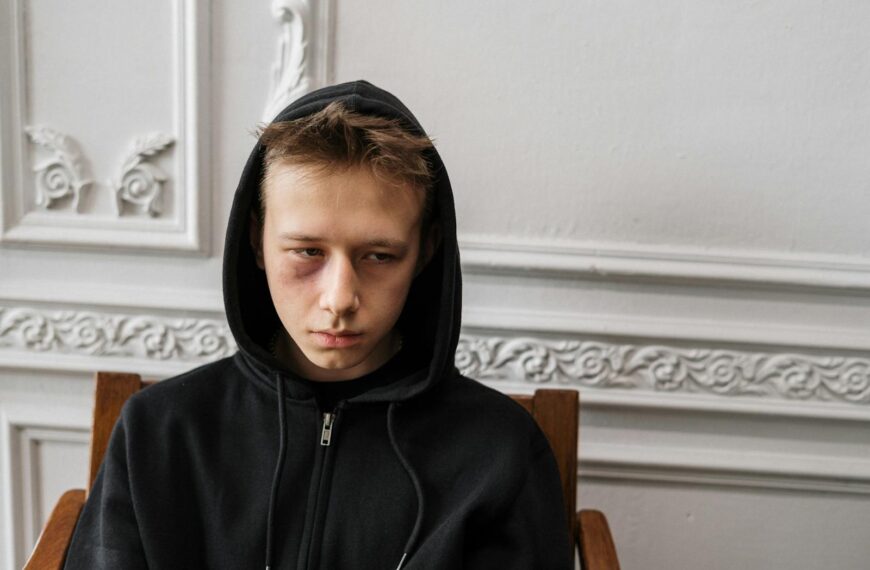 A young male sitting indoors, wearing a black hoodie with a visible bruise, showing a contemplative expression.