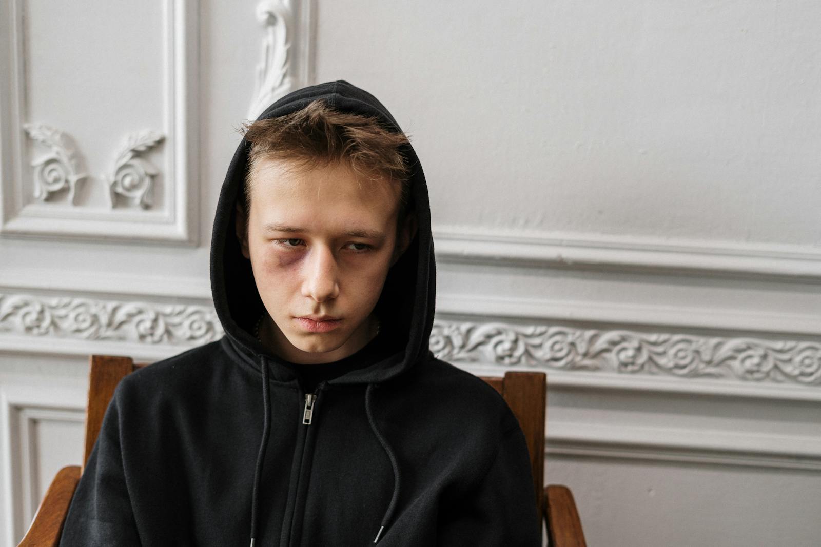 A young male sitting indoors, wearing a black hoodie with a visible bruise, showing a contemplative expression.
