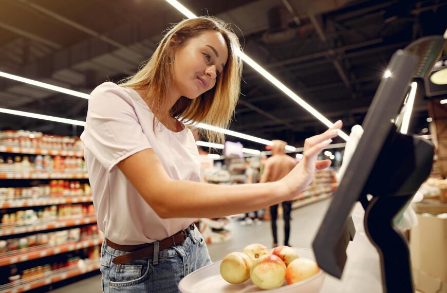 Side view of young woman in trendy clothes weighing peaches on scales while shopping in supermarket during purchase food