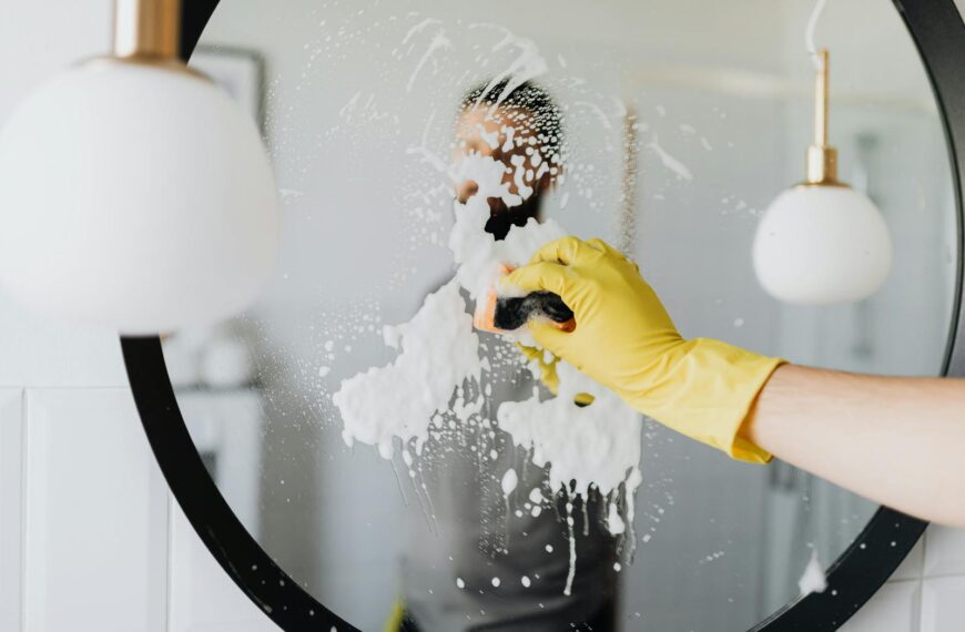 Anonymous male housekeeper in yellow rubber gloves wiping foam from round mirror in bathroom
