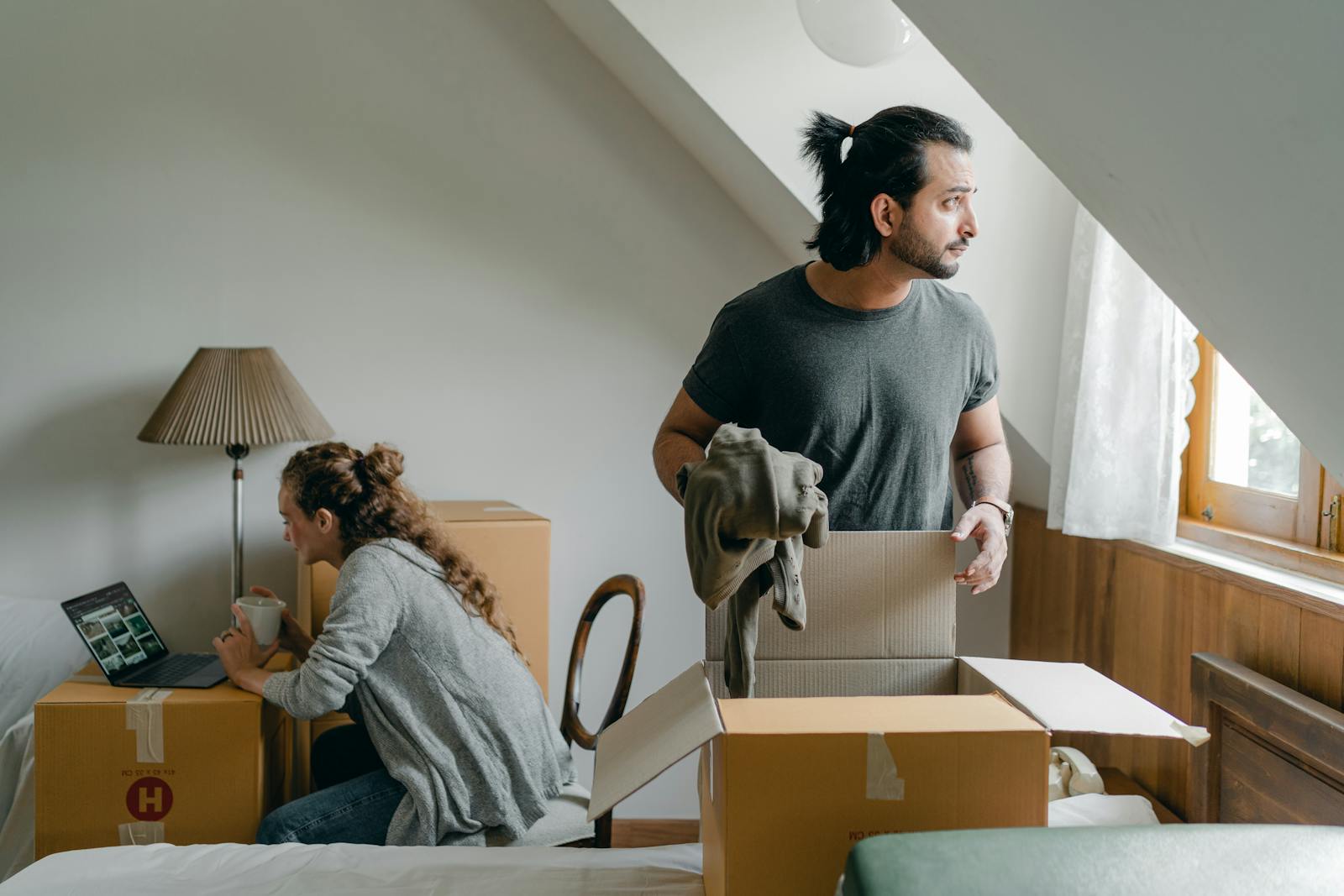 A couple unpacks boxes in a cozy attic bedroom, settling into their new home.