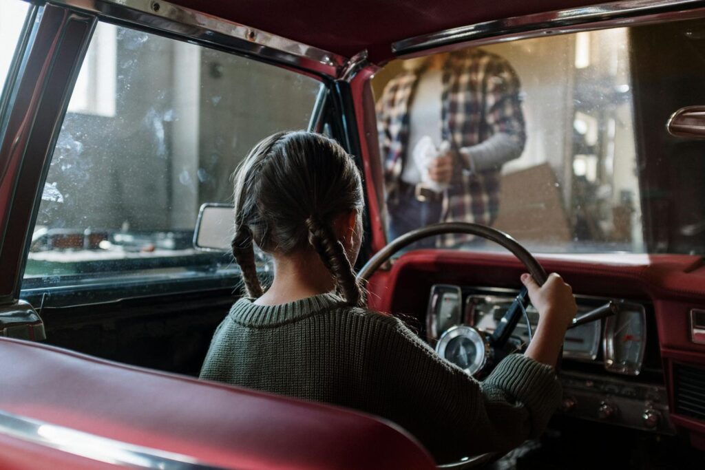 Back view of a young girl with pigtails in a vintage car interior, pretending to drive.