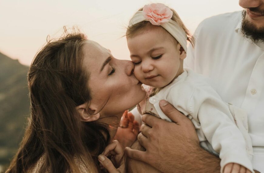 Loving mother kissing cute baby daughter held by crop father while having family day in nature