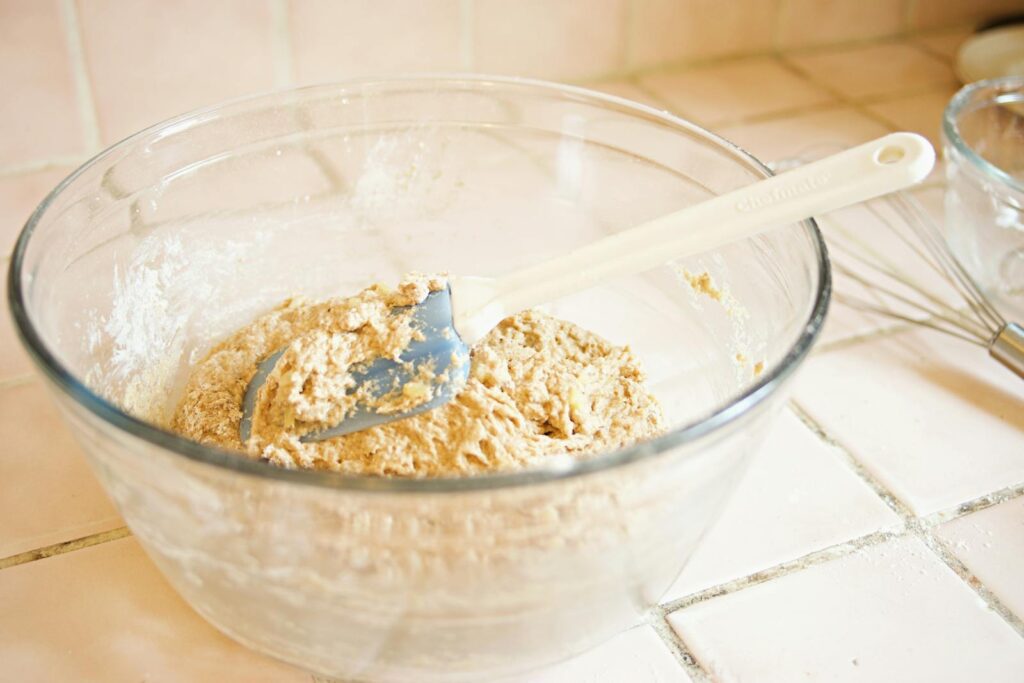 A close-up image of dough being mixed in a glass bowl with a white spatula on a tiled surface.