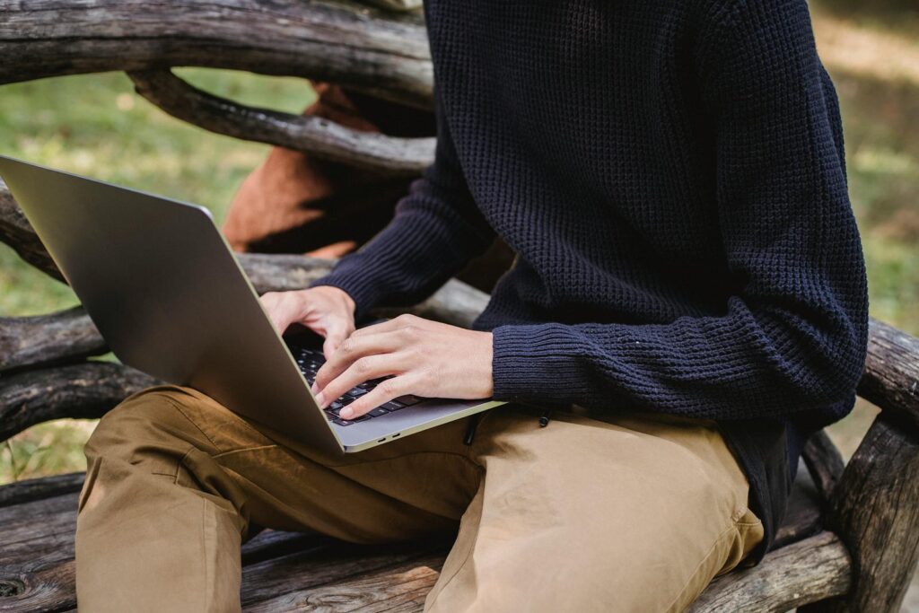 Crop anonymous male in casual clothes working on modern netbook while sitting on creative wooden bench in park