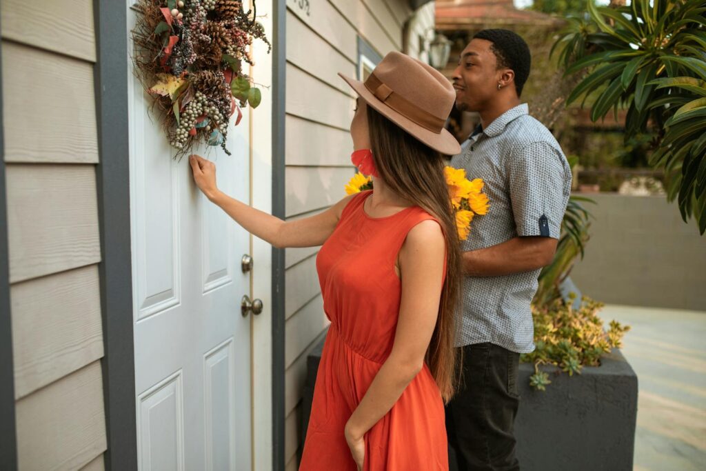 A man and a woman knocking on a door decorated with a wreath, holding flowers.
