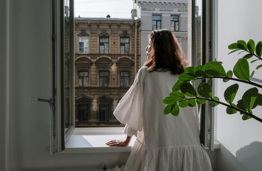 Woman in a white dress looking out an open window at classic architecture on a sunny day.