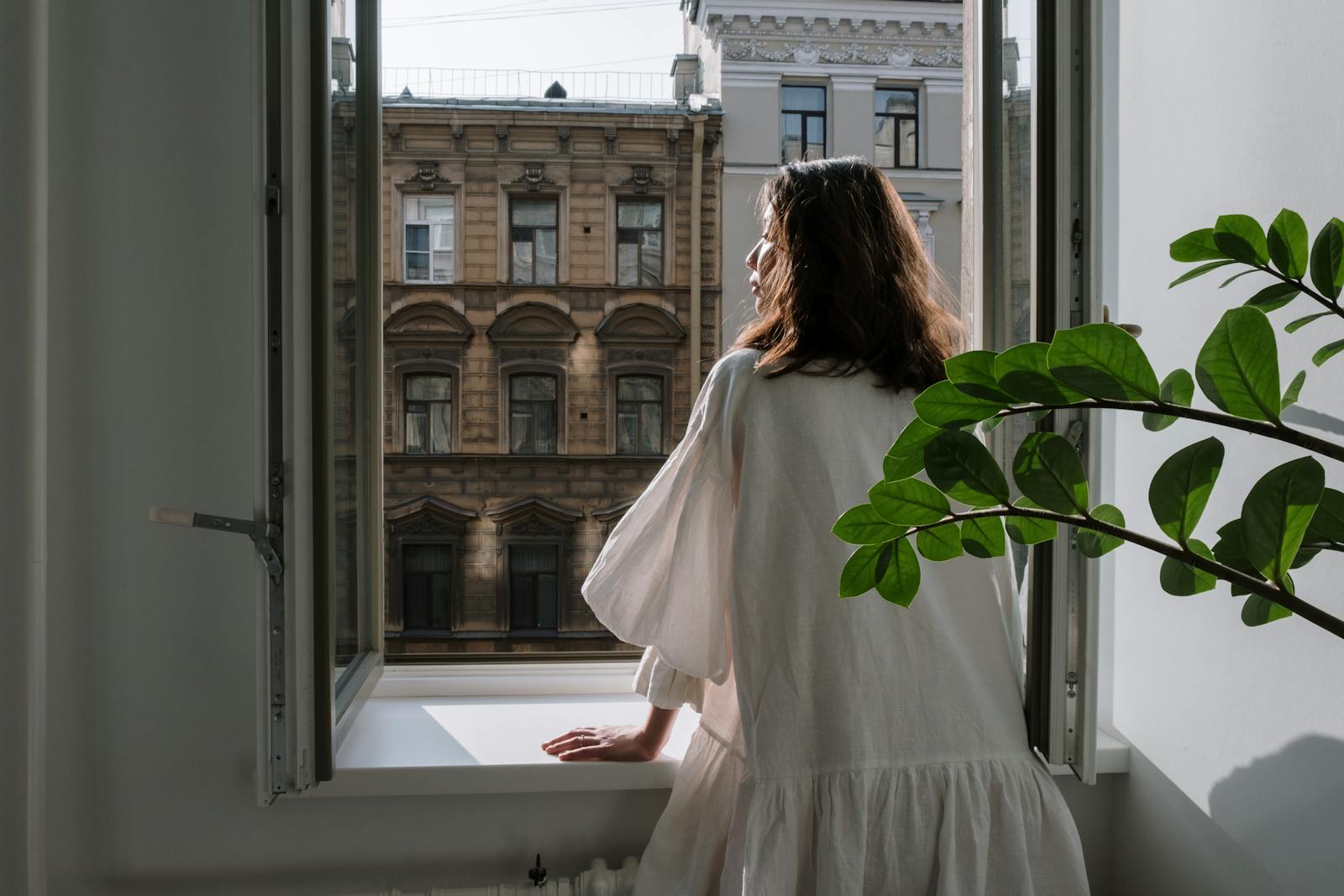Woman in a white dress looking out an open window at classic architecture on a sunny day.