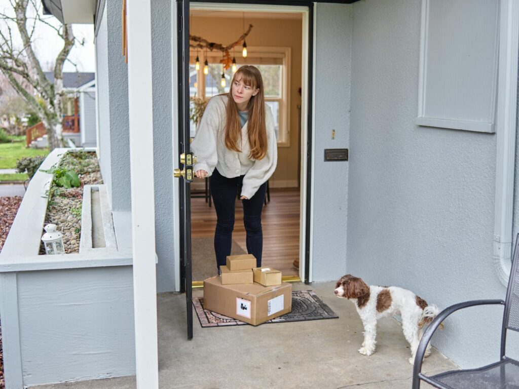 Woman opens front door to receive packages delivered on porch. Dog observes.