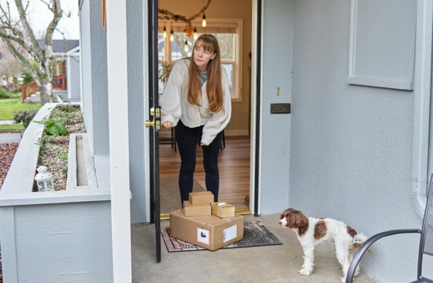 Woman opens front door to receive packages delivered on porch. Dog observes.