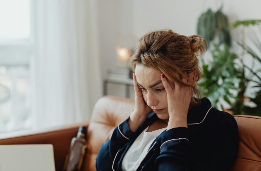 A woman in pajamas sits indoors with hands on head, showing stress and frustration. Perfect for mental health themes.