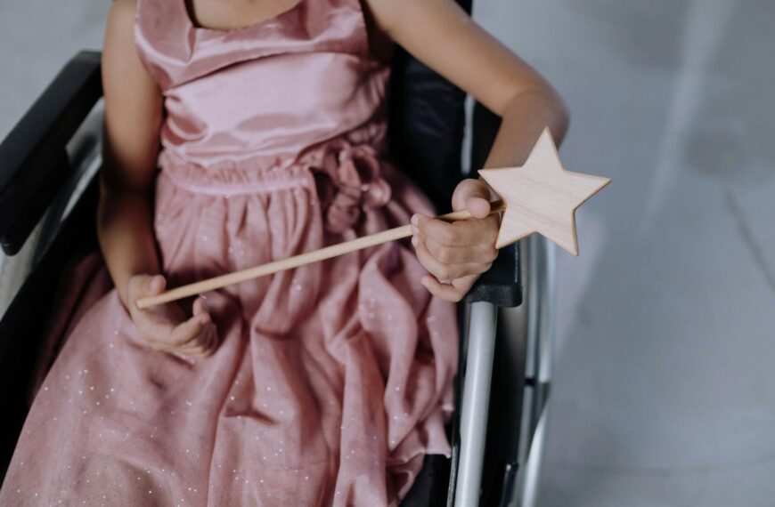 A child in a pink dress holding a star-shaped magic wand, seated in a wheelchair.