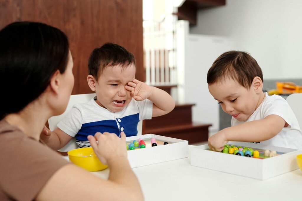 A mother attending to her crying child while another child plays contentedly indoors.