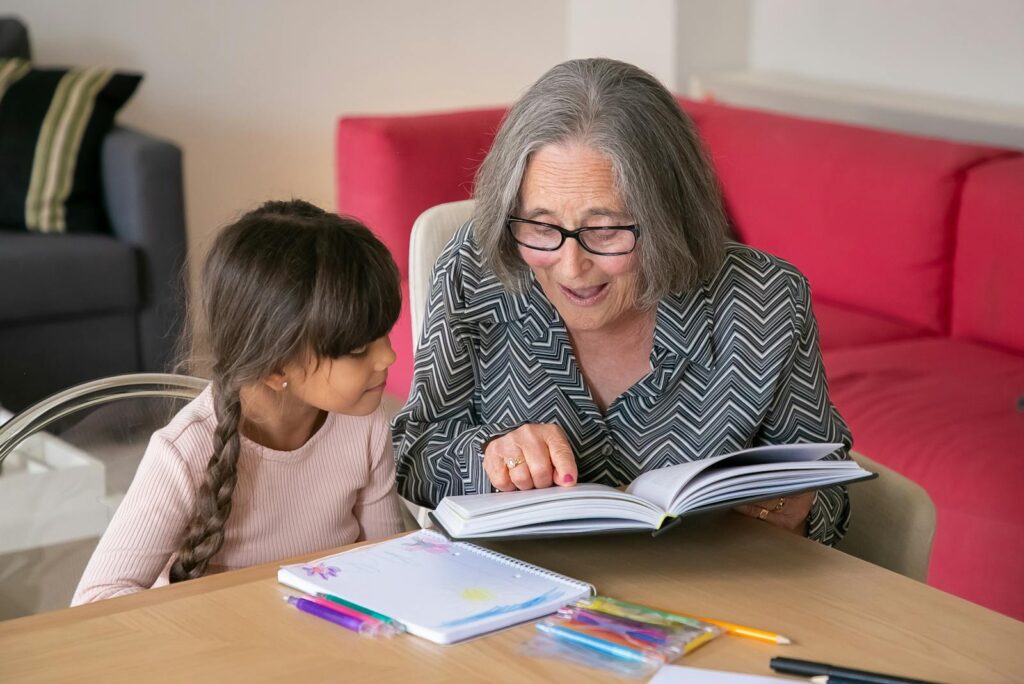 A joyful grandmother reading a book with her granddaughter indoors, bonding over learning.