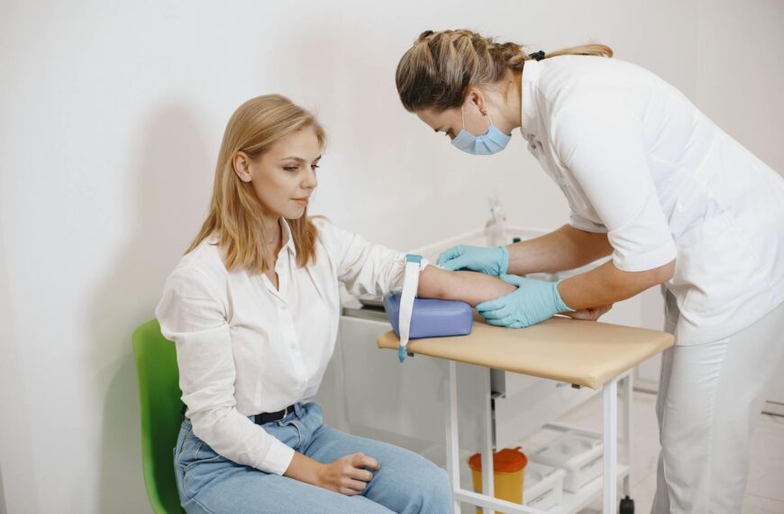 A nurse performs a blood test on a seated female patient in a medical setting.