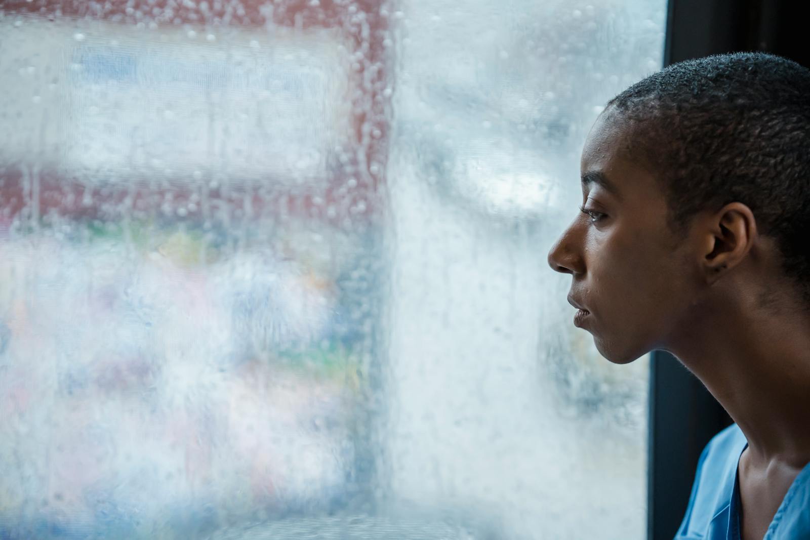 A thoughtful woman gazes out a rain-covered window, reflecting a calm mood.