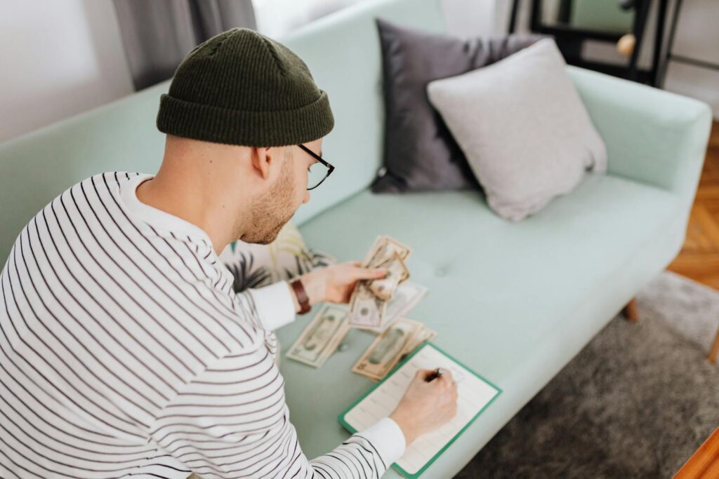 Caucasian man counting money and writing notes while sitting indoors on a couch.