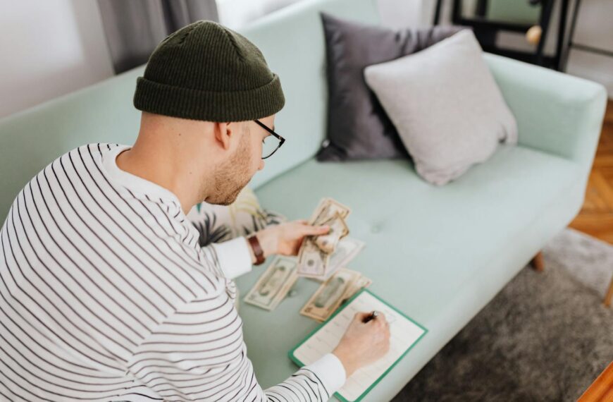 Caucasian man counting money and writing notes while sitting indoors on a couch.