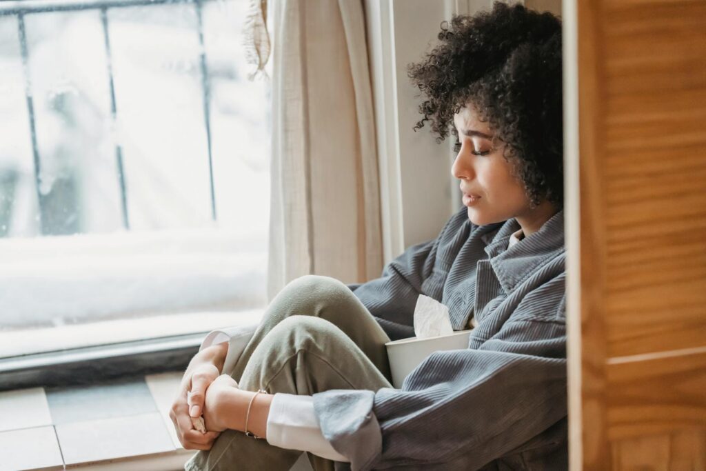 A young woman with curly hair sits alone by a window, lost in thought.
