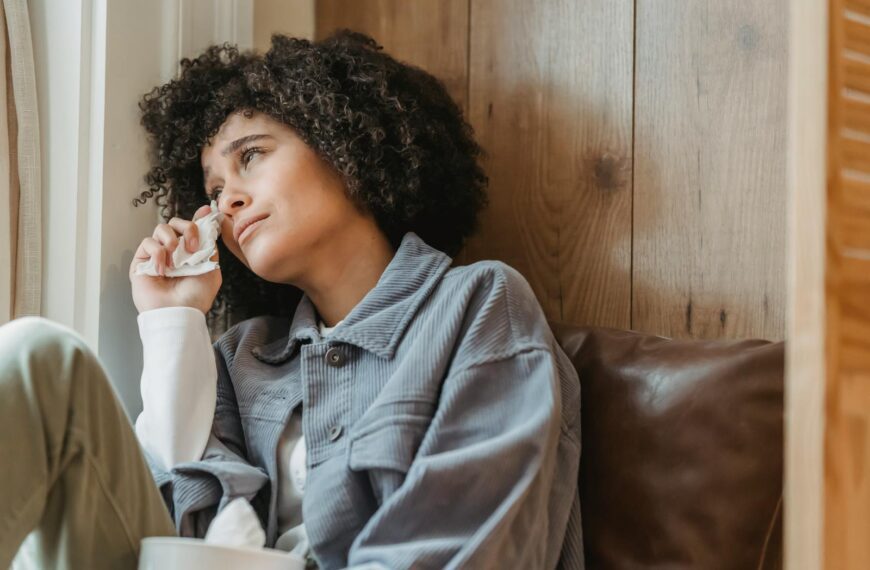 Desperate African American female with curly hair wiping face with tissue while crying from sadness on windowsill and looking away