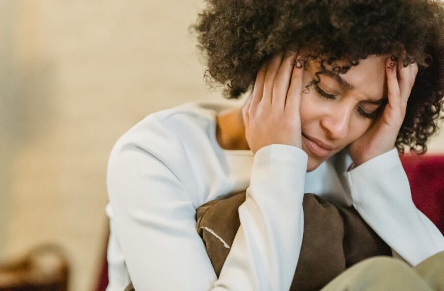 A woman with curly hair sits indoors, appearing stressed and holding her head in her hands.