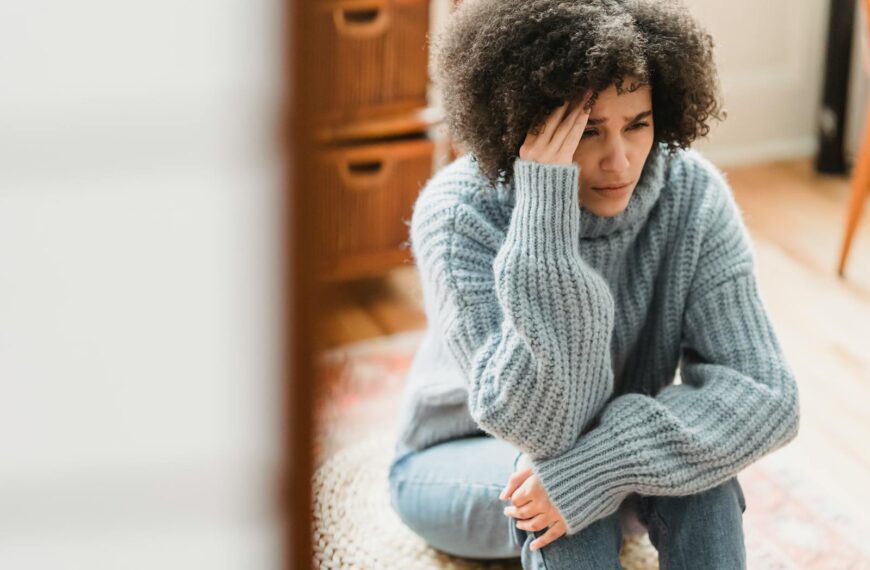 Young ethnic female in casual clothes sitting on pouf while crying with hand on head in light flat