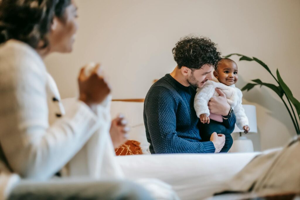 From below side view of African American mother with clothes looking at ethnic father sitting on bed while playing with baby
