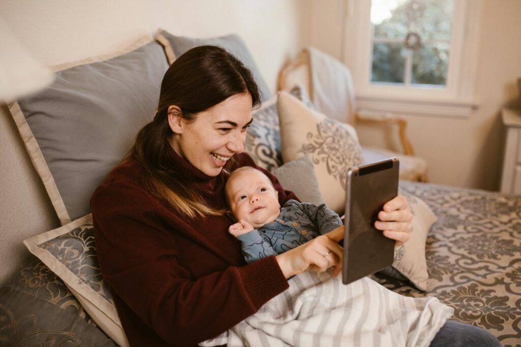 A joyful mother and her baby video calling family members using a tablet while relaxing on a cozy bed at home.