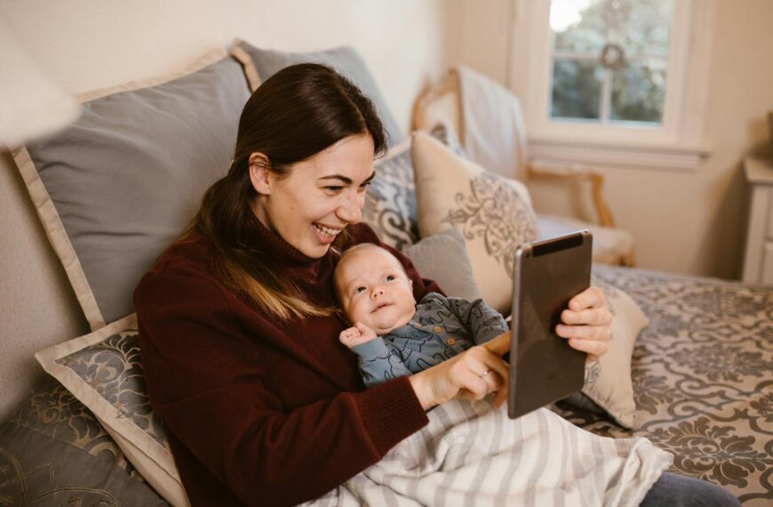 A joyful mother and her baby video calling family members using a tablet while relaxing on a cozy bed at home.