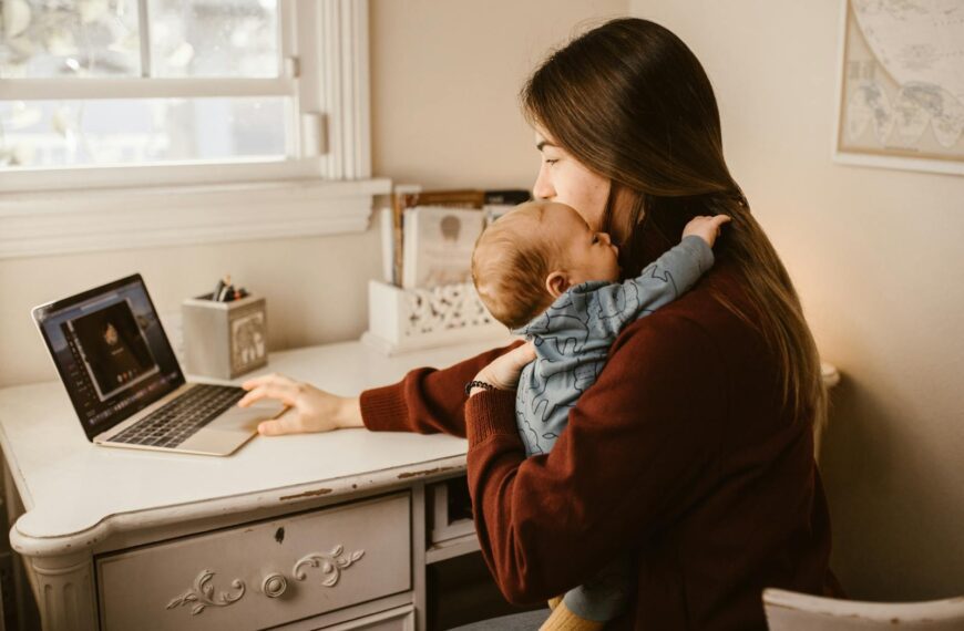 A mother multitasks with a baby in her arms while working on a laptop at home.