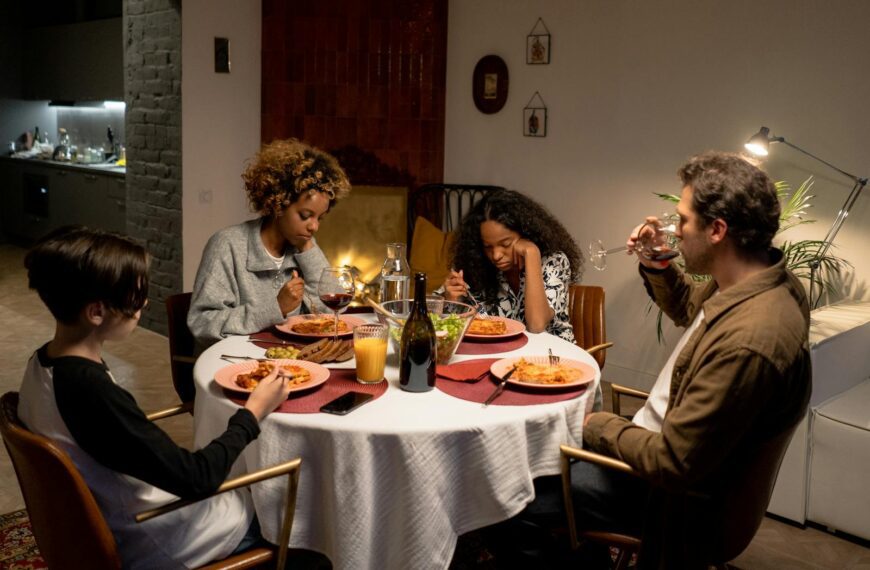 A diverse family shares a dinner of pasta and wine around a cozy indoor table setting.
