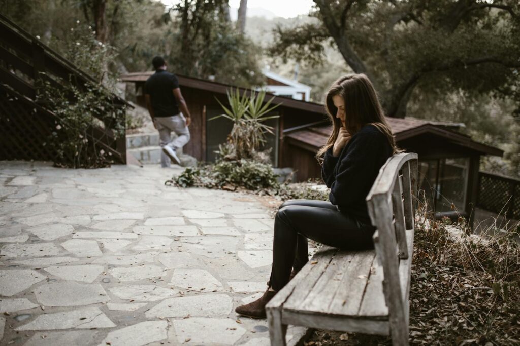 A woman sits pensively on a bench outside after a breakup, while a man walks away.