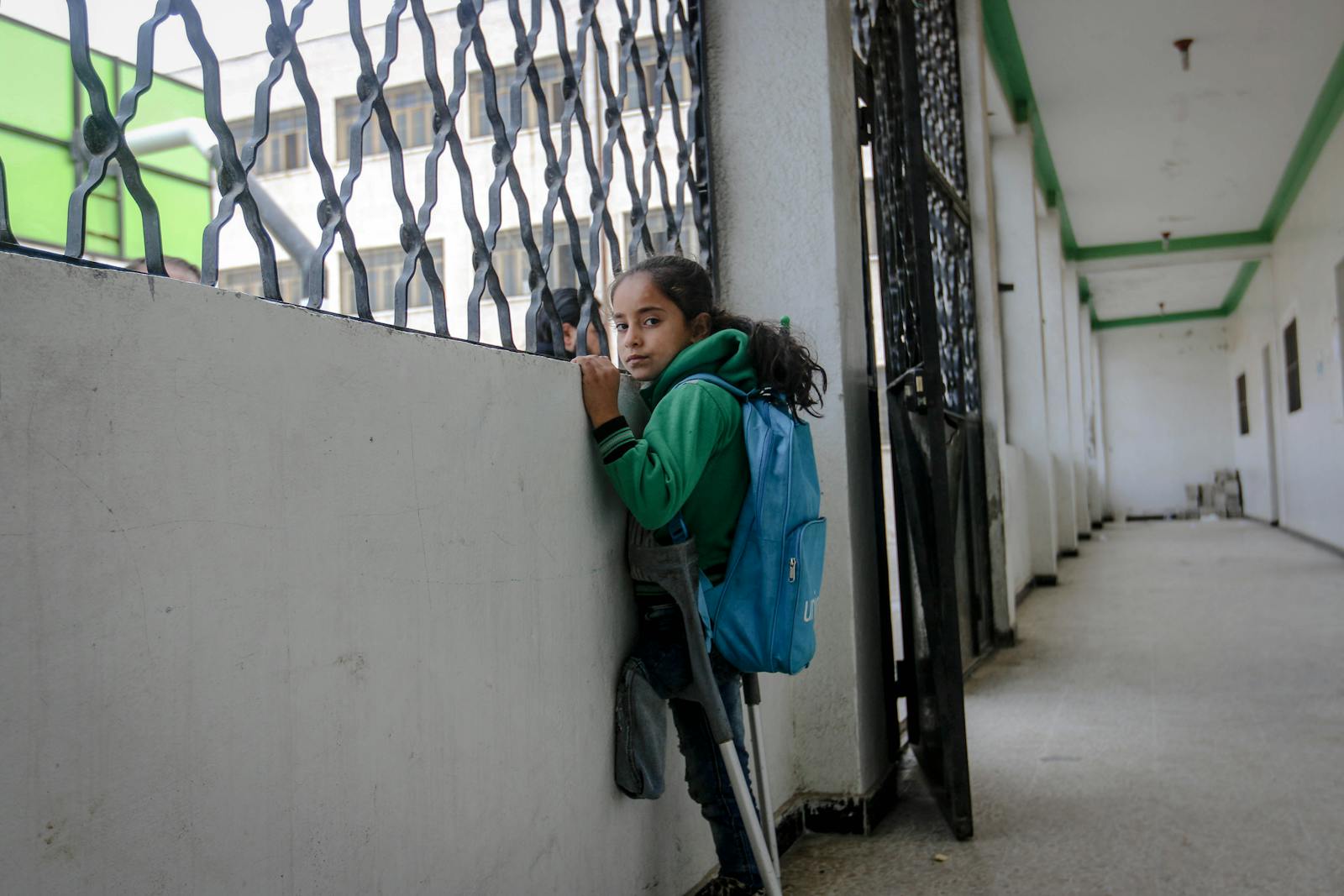 A young girl with crutches and a backpack in a corridor in Syria, looking thoughtful.