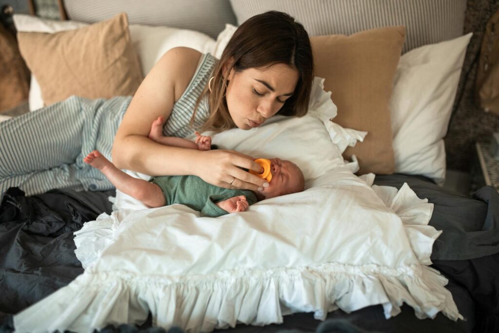 A mother gently soothes her newborn baby with a pacifier while lying in a cozy bedroom setting.