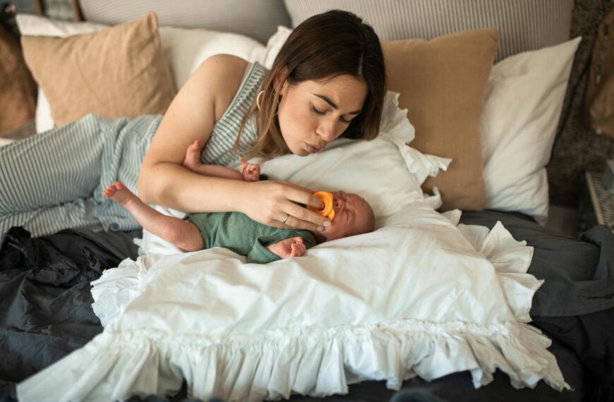 A mother gently soothes her newborn baby with a pacifier while lying in a cozy bedroom setting.