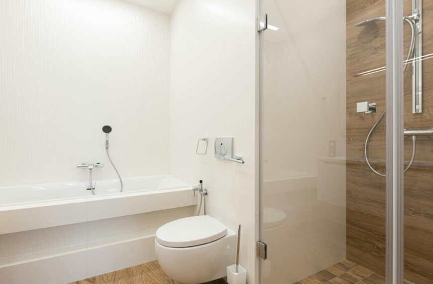 Shower cabin with glass door and ceramic bathtub and toilet bowl in spacious light bathroom with wooden floor