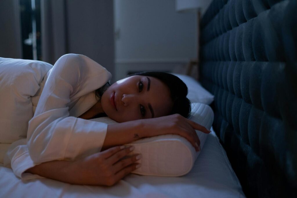 Dark bedroom scene of an Asian woman with insomnia lying on her side, looking thoughtful.