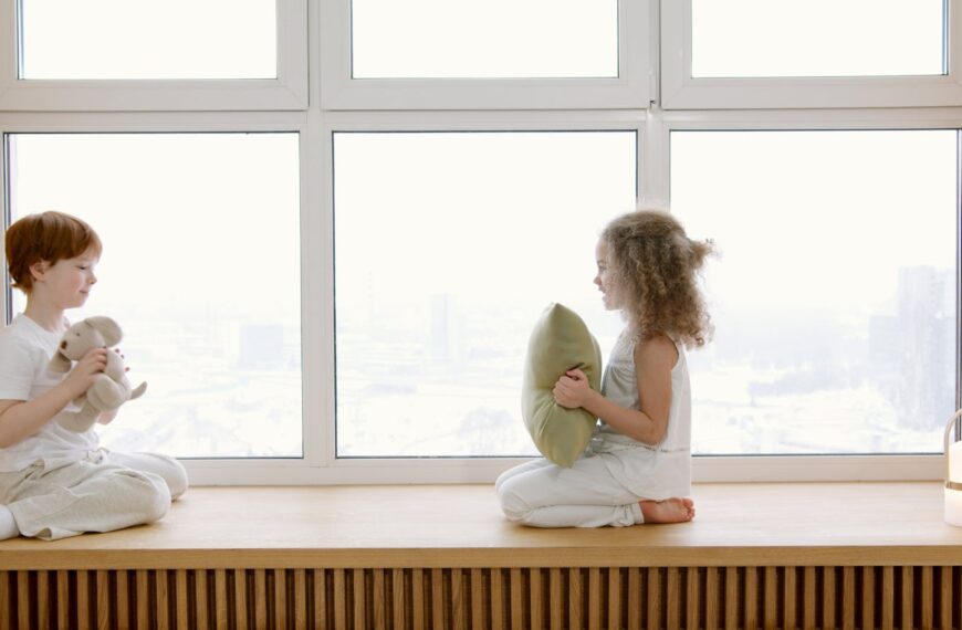 Two children sitting on a windowsill, interacting joyfully in a bright room.