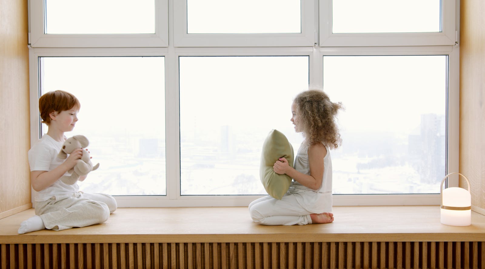 Two children sitting on a windowsill, interacting joyfully in a bright room.