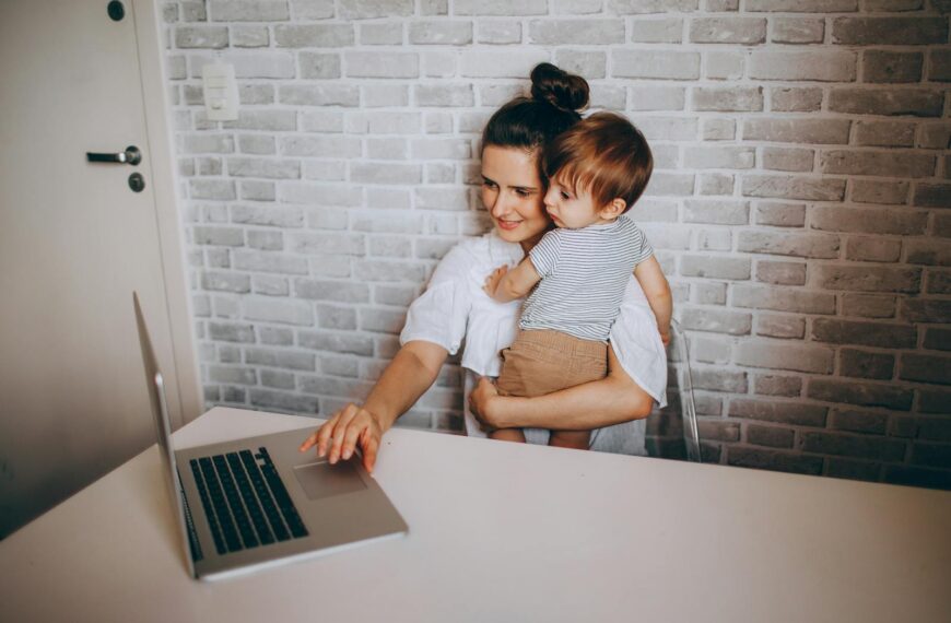 A mother works remotely on a laptop while holding her toddler in a cozy home setting.