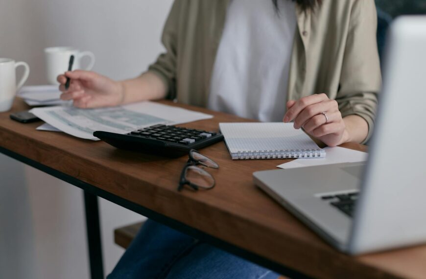 A woman manages finances at home, using a laptop and calculator on a wooden desk.