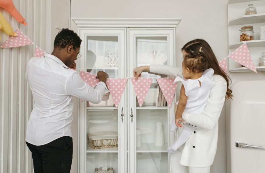 Parents setting up pink bunting for baby's birthday celebration indoors.