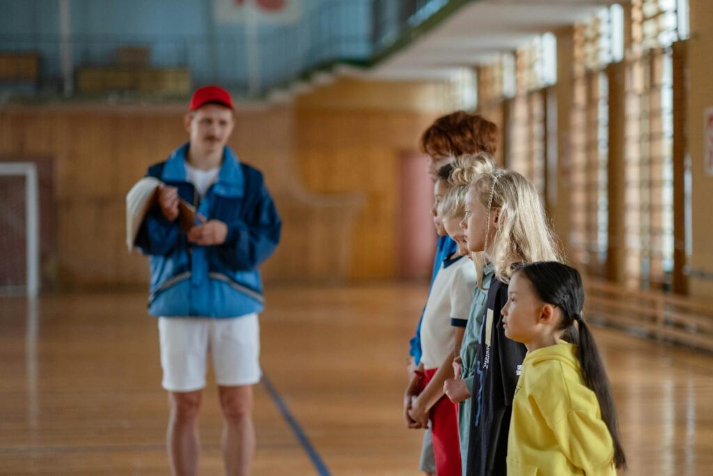 Diverse group of children standing indoors in a gym with a coach in focus.