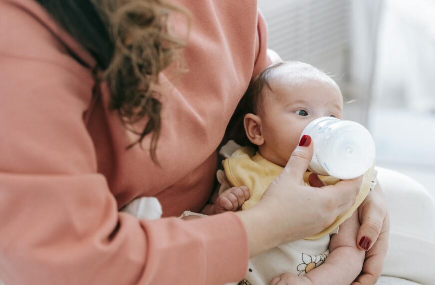 Crop anonymous mother in casual clothes feeding adorable newborn baby with bottle while sitting on sofa in light room at home