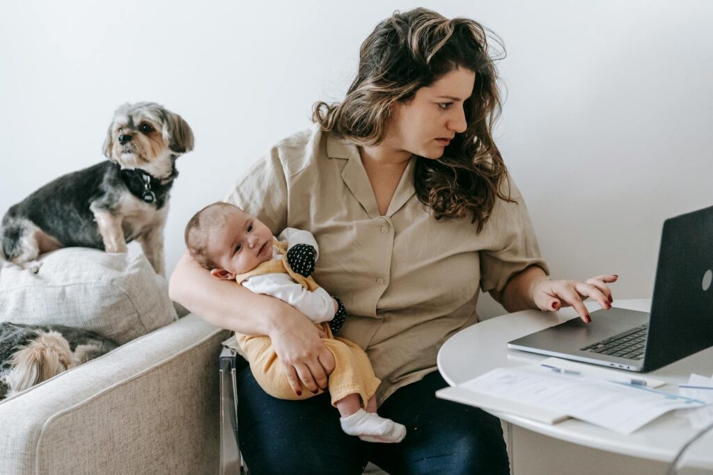 Mother balancing work from home with parenting, holding baby and typing on laptop.