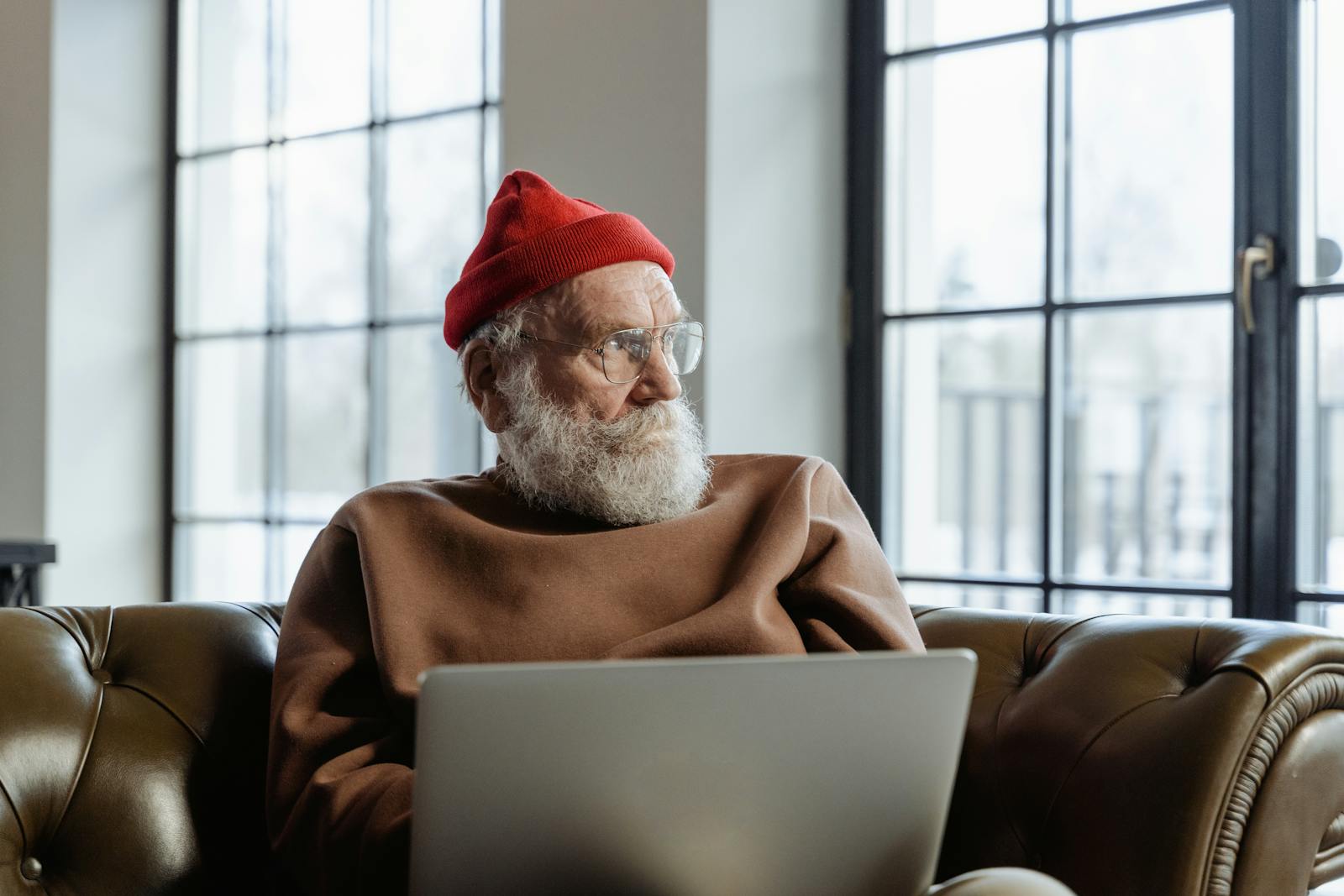 An elderly man in a red beanie is using a laptop indoors, sitting comfortably on a couch.