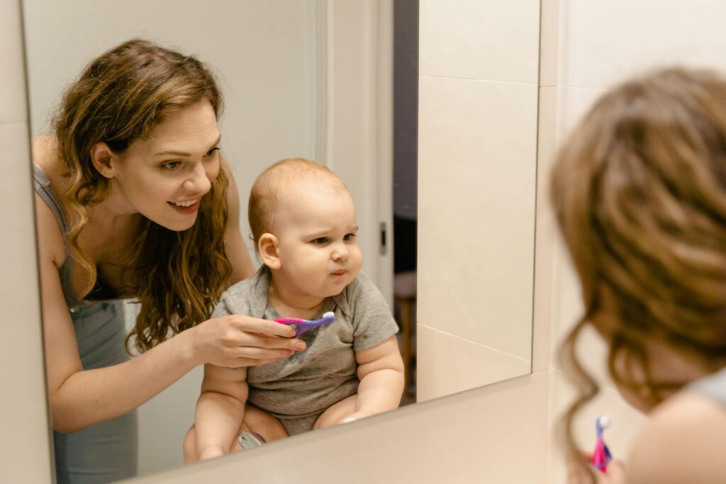 A mother helps her baby brush teeth while they share a joyful moment in front of a bathroom mirror.