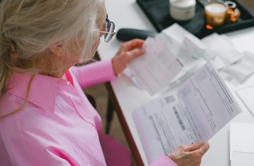 An elderly woman in glasses holds and reads important papers at a table indoors.