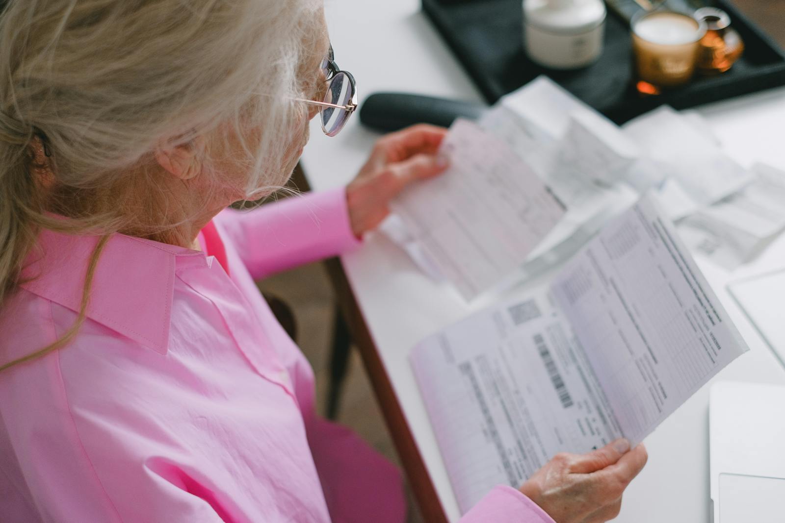 An elderly woman in glasses holds and reads important papers at a table indoors.